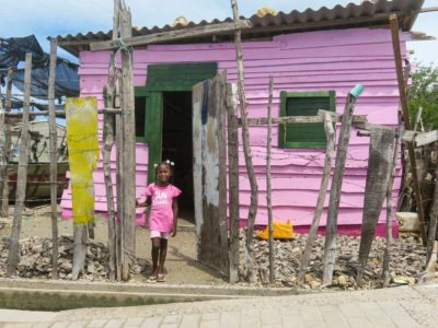 Deneilis in Front Of Her Pink Home A girl wearing pink stands outside a small pink home with a tin roof. There is a fence made of pieces of wood and barbed wire around the home.